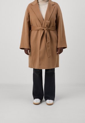 Henkilö, joka pukeutunut polvipituiseen kamelinväriseen takkiin, jossa on vyö vyötäröllä, mustiin leveälahkeisiin housuihin, valkoisiin lenkkareihin ja beigeen pooloneuleeseen.