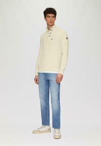 Lichtgekleurde pullover met een hoge kraag, drie knopen en een borstzak, gecombineerd met lichtblauwe jeans en grijs-witte sneakers.
