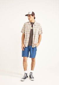 Camisa de cuadros de manga corta en beige y azul, combinada con pantalones cortos de mezclilla azules y zapatillas negras. Calcetines blancos visibles, gorra de béisbol en la cabeza.