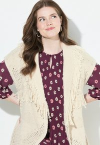 Strickweste in Creme mit Fransenverzierung über einer blauen Bluse mit Blumenmuster. Die Weste hat eine offene Vorderseite und ein strukturiertes Strickdesign.