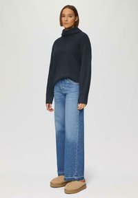Navy coltrui, ribbeltextuur, losse pasvorm. Lichtblauwe wijde jeans, vervaagd aan de zoom. Tan instappers met subtiele textuur.
