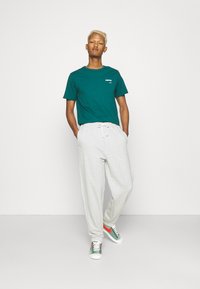 Teal t-shirt met wit logo, gecombineerd met lichtgrijze joggers. Sneakers hebben groene en rode accenten, wat een sportieve, casual look laat zien.