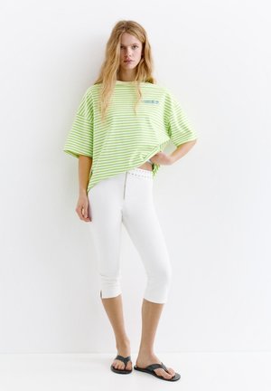 Jeune femme debout portant un t-shirt oversize rayé vert clair, un pantalon court blanc et des tongs noires sur fond blanc.