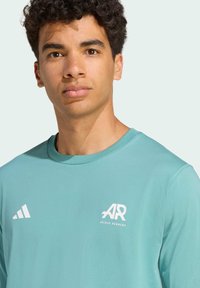T-shirt de sport turquoise clair en tissu doux, avec un col rond, logo Adidas blanc et marque "AR" sur la poitrine.