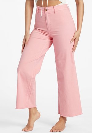 Kobieta w różowych dżinsach z wysokim stanem i szerokimi nogawkami z postrzępionymi brzegami, stoi boso z rękami spoczywającymi na udach na białym tle.