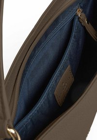 Interior de un bolso de cuero marrón con un forro de tela azul oscuro, que cuenta con un bolsillo con cremallera y una etiqueta color canela con texto en relieve.