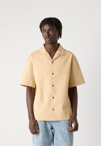Short-sleeve, light beige shirt with a textured fabric, featuring a spread collar and dark buttons, paired with light blue loose-fit jeans.