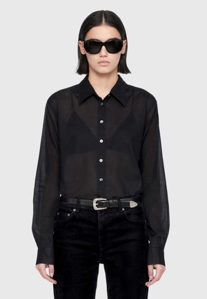 Person with straight dark hair wears large black sunglasses, a sheer black button-up shirt, black pants, and a black belt with a silver buckle.