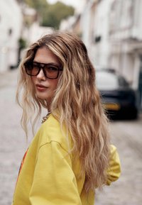 Oversized zonnebril met schildpadmontuur en gradient glazen, gedragen door een persoon in een felgele trui, met los, golvend haar.