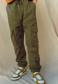 Olive green cargo pants with side pockets, elastic cuffs, and drawstring waist, paired with white and mustard sneakers. Soft, breathable fabric.