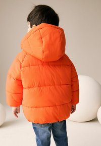 Veste orange rembourrée avec capuche, comprenant des sections matelassées, associée à un jean bleu. L'arrière-plan comprend des objets blancs, doux et ronds.