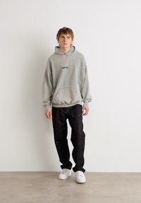 Sudadera de algodón gris con bolsillo frontal y logo, combinada con jeans de mezclilla negros y zapatillas blancas, de pie frente a un fondo liso.