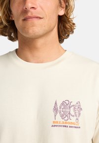 T-shirt en coton couleur crème présentant un graphisme violet de coquillages, d'un crabe et d'un poisson, avec "BILLABONG ADVENTURE DIVISION" en texte orange.
