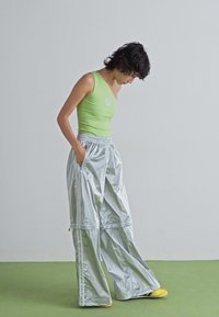 Personne aux cheveux courts et foncés portant un haut sans manches vert et un pantalon large en argent brillant, se tenant sur un sol vert contre un mur uni.