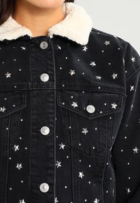 Černá džínová bunda s krémovým umělým ovčím límcem, stříbrnými hvězdicovými ozdobami a heslovými knoflíky. Obsahuje dvě přední kapsy.