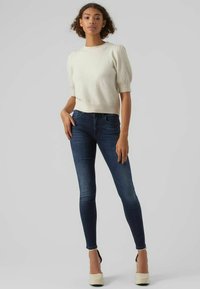 Ljust beige stickat tröja med korta puffärmar, tillsammans med mörkblå skinny jeans och krämfärgade plattformsskor. Slät textur genom hela plagget.