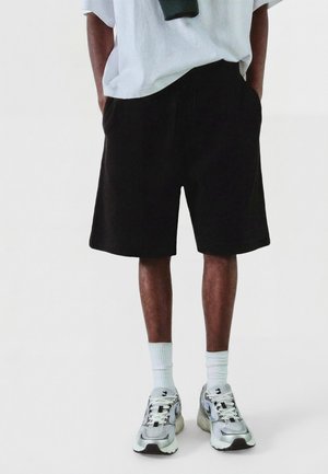 Personne portant un short noir, des chaussettes blanches montantes, des baskets grises et blanches et une chemise blanche avec les mains dans les poches.