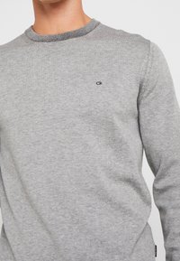 Gray crewneck sweater made of soft knit fabric, featuring a ribbed neckline and a small black logo on the chest. Smooth texture and fitted shape.