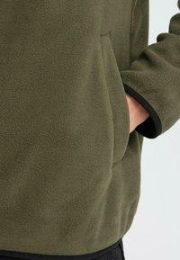 Kergedest fleece-jakk muted roheline tooni, pehme tekstuuriga, mustadeäärtega varrukatel ja paremal küljel esikotis.