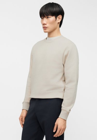 Μπεζ φούτερ με στρογγυλή λαιμόκοψη, μακριά μανίκια και ριπ μανσέτες. Απαλή υφή, μινιμαλιστικός σχεδιασμός και διακριτικές λεπτομέρειες λογότυπου στο μπροστινό μέρος.