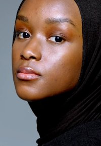 Svart hijab draperer over en jevn, lys brun hudtone. Modellens subtile sminke fremhever hennes naturlige trekk mot en nøytral bakgrunn.