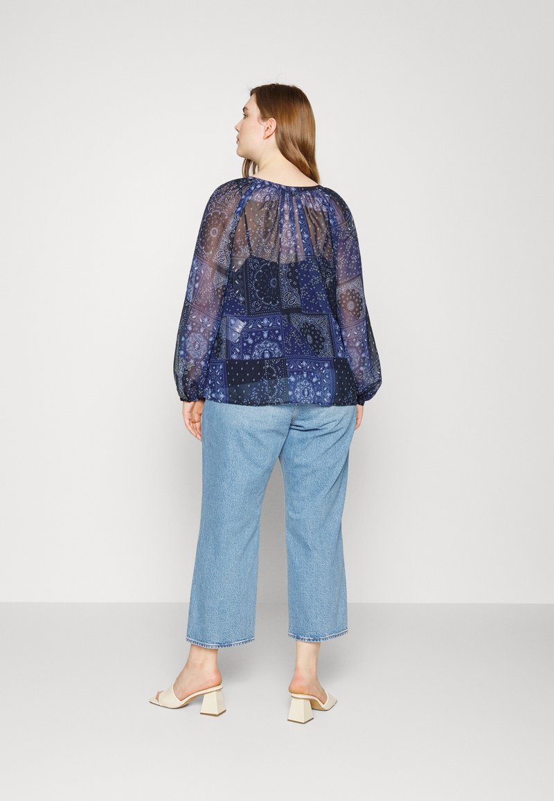 Durchsichtige marineblaue Bluse mit Ballonärmeln, gemustertem Stoff und gerafftem Ausschnitt; kombiniert mit hellblauen, geraden Jeans und cremfarbenen Blockabsatzsandalen.