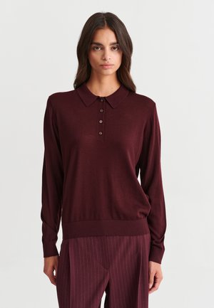 Femme aux longs cheveux foncés portant un polo à manches longues bordeaux foncé et un pantalon rayé assorti, debout devant un fond clair uni.