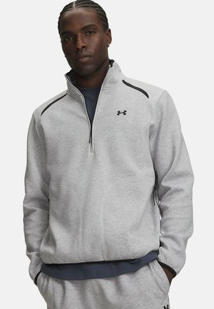 Man met gevlochten haar, die een lichtgrijs sportjack met rits en bijpassende joggingbroek draagt, staand met handen in de zakken.