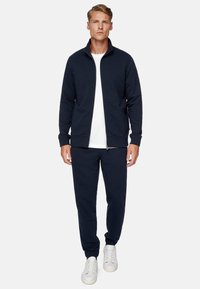 Giacca a zip blu navy e pantaloni jogger coordinati realizzati in tessuto morbido, con polsini a costine e t-shirt bianca a girocollo. Sneakers bianche completano l'outfit.
