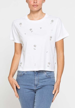 Donna che indossa una t-shirt bianca a maniche corte con piccoli ornamenti floreali argento e jeans blu a vita alta.