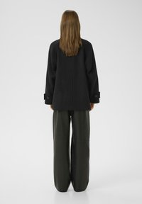 Black oversized coat with dropped shoulders, featuring buttoned cuffs and a central seam. Paired with wide-legged green pants.