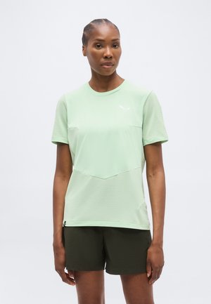 Nő világoszöld, rövid ujjú sportpólóban és sötétzöld rövidnadrágban, egyszínű fehér háttér előtt áll.