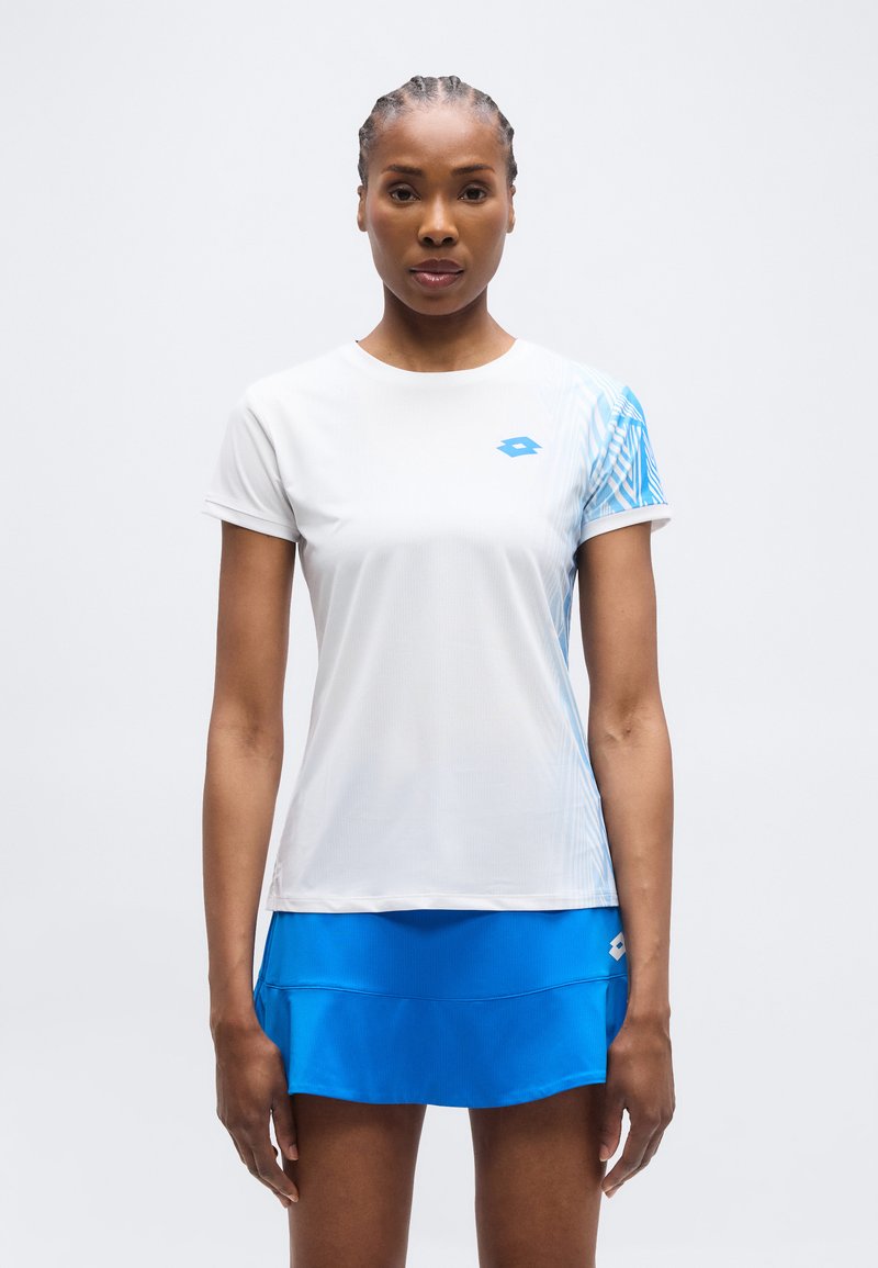 Γυναίκα που στέκεται με το πρόσωπο προς τα εμπρός, φορώντας λευκό αθλητικό πουκάμισο με μπλε μοτίβα και φωτεινή μπλε αθλητική φούστα.