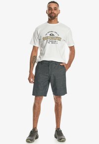 Herr T-shirt från Quiksilver, vit med svart och gul logotyp, bärs med mörka, strukturerade shorts. Sneakers har en grå färg.