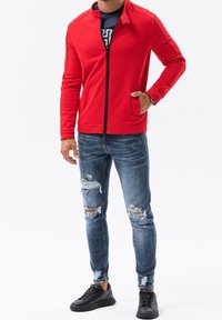 Giacca rossa con zip e colletto alto, in tessuto strutturato, abbinata a jeans blu scoloriti e sneakers nere. Look casual e moderno.