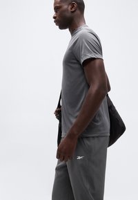 Άνδρας με γκρι αθλητική ένδυση κρατώντας μια μαύρη τσάντα ώμου μπροστά σε απλό ανοιχτό φόντο, πλαϊνή όψη.