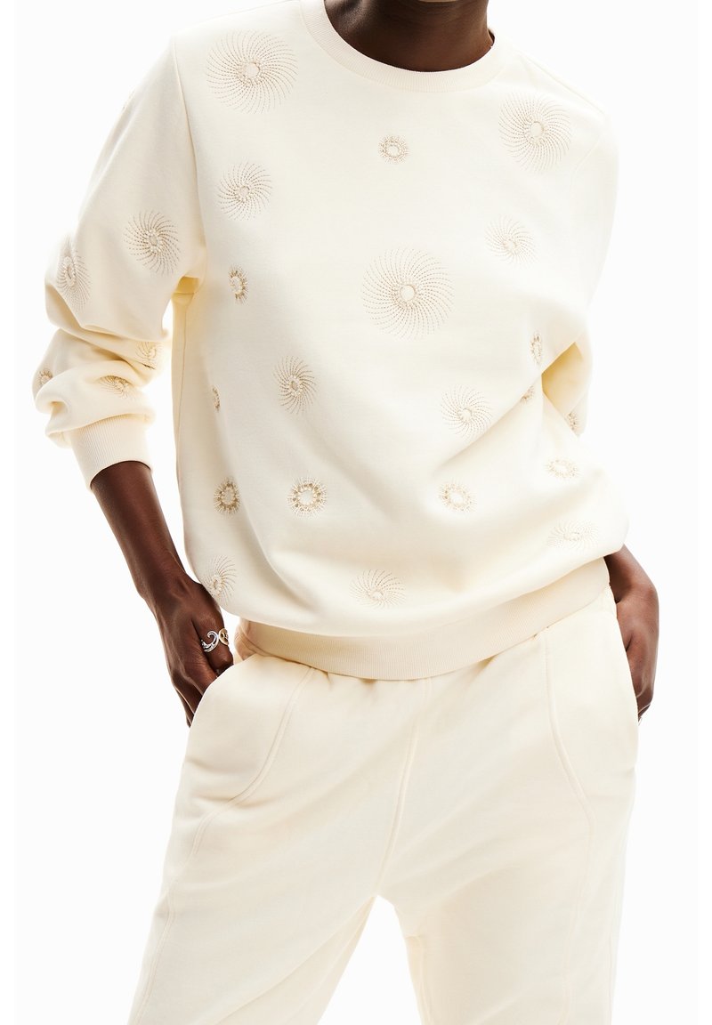 Personne portant un sweat-shirt crème avec des motifs brodés circulaires et un pantalon crème assorti, les mains dans les poches, teint de peau marron.