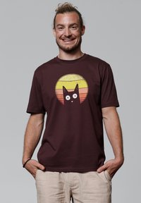 T-shirt en coton bordeaux avec un motif de chat en cartoon sur un fond de rayons jaunes et oranges. Manches courtes et col ras du cou.