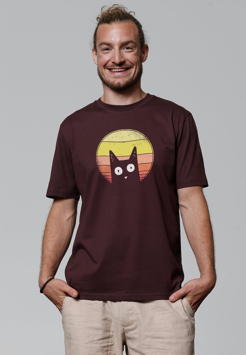 T-shirt en coton bordeaux avec un motif de chat en cartoon sur un fond de rayons jaunes et oranges. Manches courtes et col ras du cou.