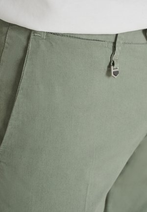 Primo piano della vita di pantaloni verde chiaro con dettaglio a clip metallica e camicia bianca infilata visibile in alto.