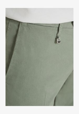 Primo piano della vita di pantaloni verde chiaro con dettaglio a clip metallica e camicia bianca infilata visibile in alto.