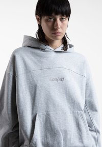 Pilkas hoodie, pagamintas iš minkštos medžiagos, su priekinėmis kišenėmis, su raišteliu reguliuojamą kapučiu ir išsiuvinėtu logotipu krūtinėje. Atsipalaidavęs siluetas, minimalus dizainas.