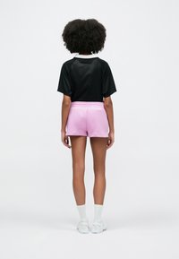 Μαύρο πουκάμισο από δίχτυ με κοντά μανίκια, συνδυασμένο με ροζ σορτς. Το μοντέλο φορά λευκές κάλτσες και παπούτσια, οπτικά από την πίσω πλευρά.