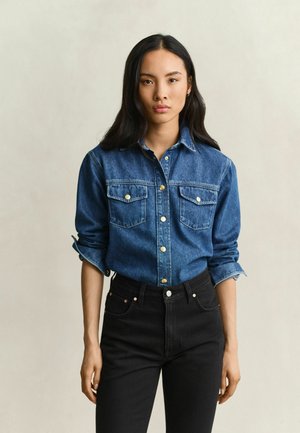 Jeune femme aux longs cheveux foncés portant une chemise en jean bleue à manches retroussées et un jean noir taille haute sur un fond uni.