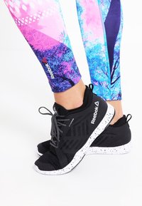 Svarta Reebok-sneakers med en fläckig vit sula, i kombination med färgglada, mönstrade leggings i rosa och blå toner.