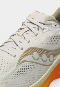 Κοντινό πλάνο λευκού παπουτσιού για τρέξιμο με διαπνέον πλέγμα, λευκά κορδόνια, μπεζ λεπτομέρειες και πορτοκαλί σόλα.