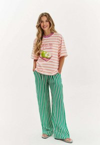 Svetloružový a biely prúžkovaný oversize tričko s grafikou zelenej jablka a textom. Voľné zelené vertikálne pruhované nohavice. Ploché sandále.