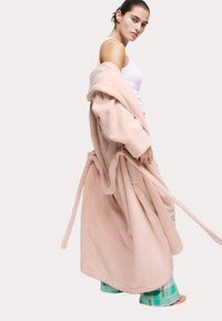 Mjukt rosa, oversized fleece-morgonrock med långa ärmar och knytbelte. Modellen bär en vit linne och rutiga pyjamasbyxor.