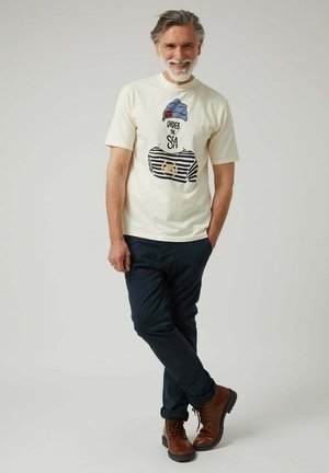 Camiseta de algodón color beige claro con un estampado gráfico de un traje de baño a rayas y un sombrero azul. Combinada con pantalones azul marino oscuros y botas marrones de cordones.