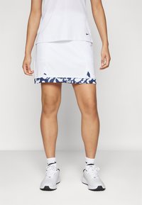 White athletic skirt with navy patterned hem, featuring side slits for mobility. Paired with a white top and white sneakers.